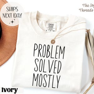 May include: Ivory-colored t-shirt with the text "PROBLEM SOLVED MOSTLY" in black. The shirt has a crew neck and short sleeves. A gold necklace with a pendant is draped on the shirt. The Ivy Threads Co. logo is in the upper right corner.