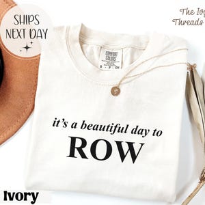 May include: Ivory-colored t-shirt with the text "it's a beautiful day to ROW" in black. A gold necklace with a pendant rests on the shirt. The shirt has a Comfort Colors tag.