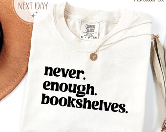 May include: Ivory-colored t-shirt with the text "never. enough. bookshelves." in bold black letters. The shirt has a crew neck and a small tag on the collar. A gold necklace with a pendant is draped across the shirt.