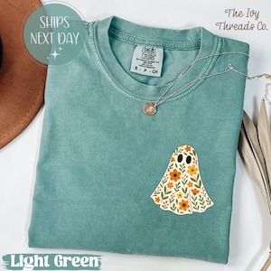 Puede incluir: Camiseta verde claro con un diseño floral de fantasma. La camiseta tiene cuello redondo y mangas cortas. La etiqueta dice "Comfort Colors". Un collar plateado con un colgante está en la camiseta. El texto "The Ivy Threads Co." está en la esquina superior derecha.