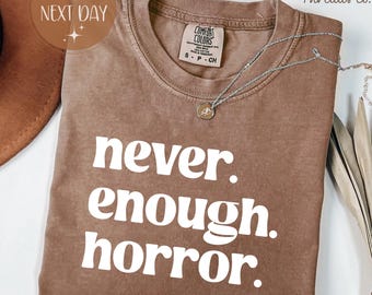 Könnte beinhalten: Espressofarbenes T-Shirt mit dem weißen Text "never. enough. horror.". Das Shirt hat einen Rundhalsausschnitt und kurze Ärmel. Eine silberne Halskette mit Anhänger liegt über dem Shirt. Das Shirt ist mit "Comfort Colors" gekennzeichnet.