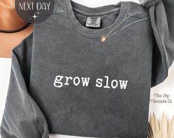 May include: A dark gray sweatshirt with the words "grow slow" in white block letters. The sweatshirt has a crew neck and long sleeves. A delicate necklace with a pendant is visible. The word "Pepper" is printed at the bottom.
