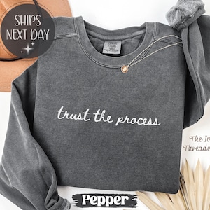 May include: A dark gray sweatshirt with the phrase "trust the process" embroidered in white. The sweatshirt has long sleeves and a crew neck. A gold necklace is draped on the front. The word "Pepper" is at the bottom.