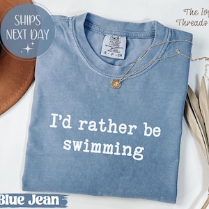 Könnte beinhalten: Ein blaues Jeans-T-Shirt mit dem weißen Text "I'd rather be swimming". Eine goldene Halskette mit Anhänger liegt auf dem Shirt. Das Shirt hat ein Comfort Colors-Etikett.