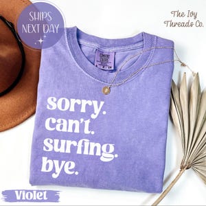 May include: A violet-colored t-shirt with the text "sorry. can't. surfing. bye." in white. The shirt has a crew neck and short sleeves. A gold necklace with a pendant is draped on the shirt. The words "SHIPS NEXT DAY" are in a circle.