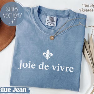 Puede incluir: Una camiseta azul vaquera Comfort Colors con la frase "joie de vivre" en blanco. Un collar dorado con un colgante descansa sobre la camisa. La camisa tambi&eacute;n presenta un dise&ntilde;o de flor de lis blanca.