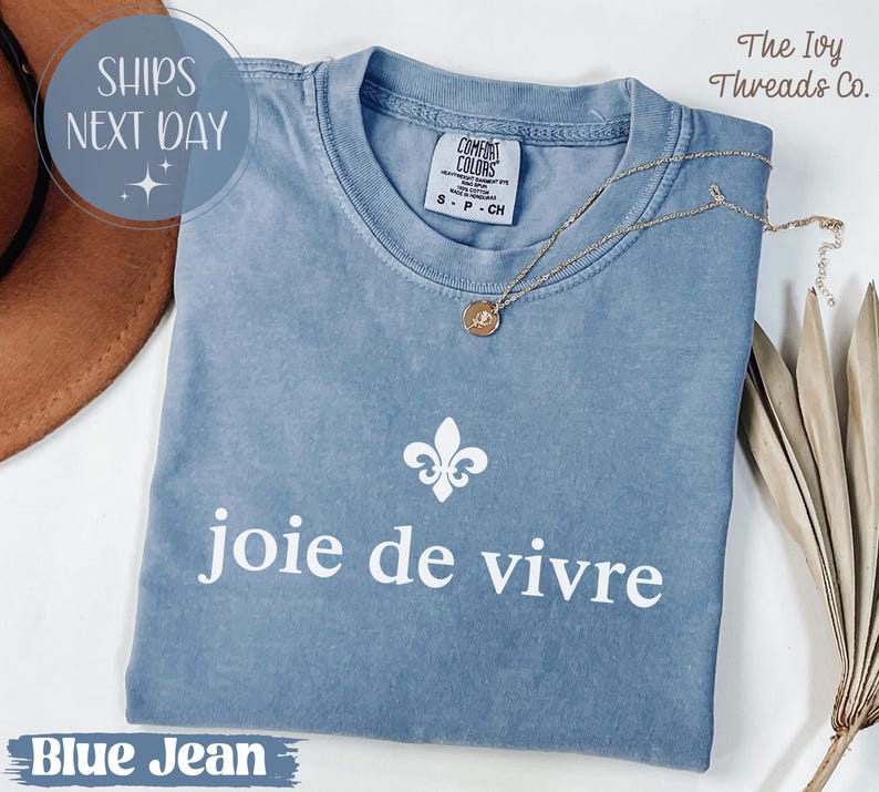 Puede incluir: Una camiseta azul vaquera Comfort Colors con la frase "joie de vivre" en blanco. Un collar dorado con un colgante descansa sobre la camisa. La camisa tambi&eacute;n presenta un dise&ntilde;o de flor de lis blanca.