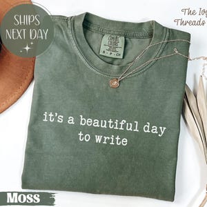 May include: Moss green t-shirt with the text "it's a beautiful day to write" in white. A gold necklace with a pendant rests on the shirt. The shirt is folded and displayed with a hat and dried plant.