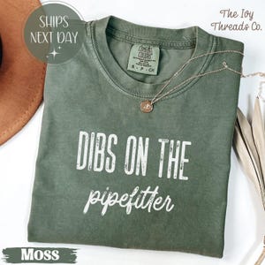 May include: A moss green t-shirt with the text "DIBS ON THE pipefitter" in white. The shirt has a crew neck and short sleeves. A gold necklace with a pendant is draped on the shirt. The shirt is from The Ivy Threads Co.