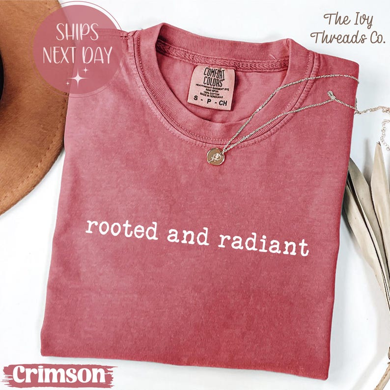 May include: Crimson-colored t-shirt with the text "rooted and radiant" in white. The shirt has a crew neck and a small tag on the collar. A silver necklace with a pendant is draped on the shirt.