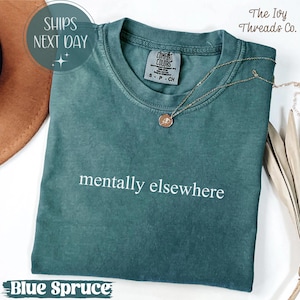 May include: A blue spruce Comfort Colors t-shirt with the phrase "mentally elsewhere" in white. The shirt has a crew neck and short sleeves. A gold necklace with a pendant rests on the shirt. The text "The Ivy Threads Co." is visible.