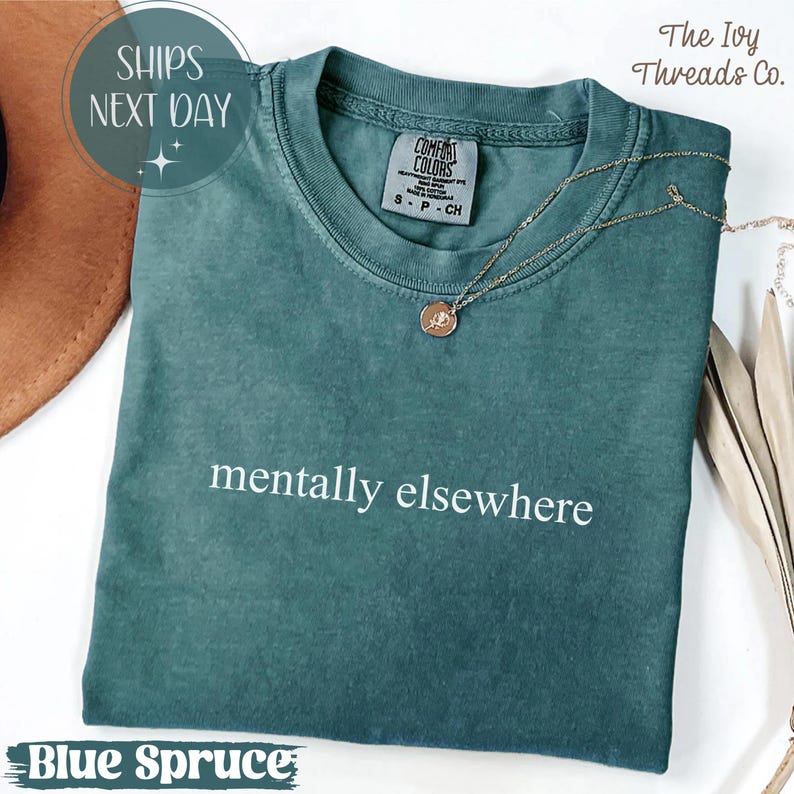 May include: A blue spruce Comfort Colors t-shirt with the phrase "mentally elsewhere" in white. The shirt has a crew neck and short sleeves. A gold necklace with a pendant rests on the shirt. The text "The Ivy Threads Co." is visible.