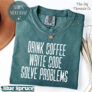Peut inclure: T-shirt bleu &eacute;pic&eacute;a avec le texte blanc "DRINK COFFEE WRITE CODE SOLVE PROBLEMS". Le t-shirt a un col rond et des manches courtes. Un collier en or avec un pendentif est drap&eacute; sur le t-shirt.