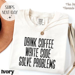Peut inclure: T-shirt ivoire avec l'inscription noire "DRINK COFFEE WRITE CODE SOLVE PROBLEMS". Le t-shirt a un col rond et des manches courtes. Un collier dor&eacute; avec un pendentif est drap&eacute; sur le t-shirt. Les mots "SHIPS NEXT DAY" sont dans un cercle.