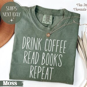 May include: Moss green t-shirt with the text "DRINK COFFEE READ BOOKS REPEAT" in white. The shirt has a crew neck and short sleeves. A gold necklace with a pendant rests on the shirt.