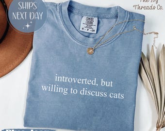 Puede incluir: Una camiseta Comfort Colors azul vaquero con el texto "introverted, but willing to discuss cats" en blanco. Un collar dorado con un colgante descansa sobre la camisa.