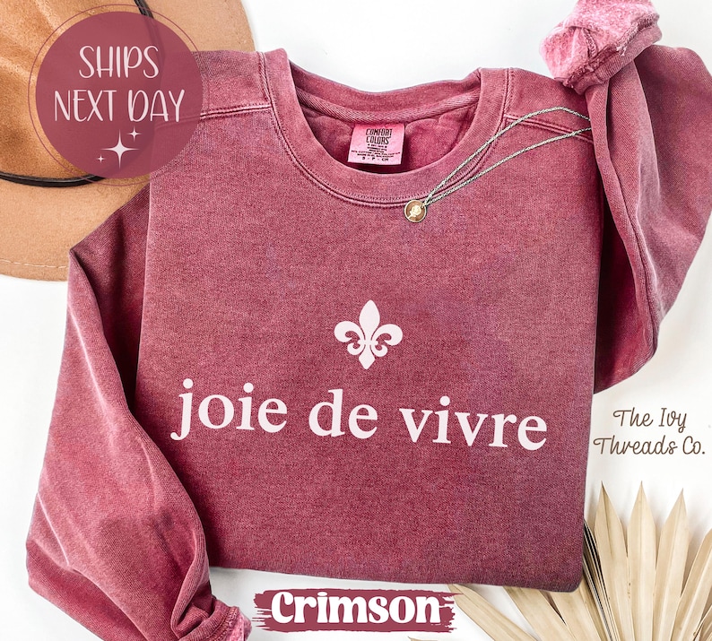 Puede incluir: Sudadera de color carmes&iacute; con la frase "joie de vivre" en escritura blanca. Un collar plateado con un peque&ntilde;o colgante descansa en la parte delantera. La sudadera presenta un s&iacute;mbolo de flor de lis y las palabras "The Ivy Threads Co."