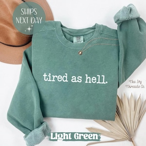 May include: Light green sweatshirt with the phrase "tired as hell" in white lettering. The sweatshirt has a crew neck and long sleeves with rolled cuffs. A gold necklace with a small pendant is visible. The words "Light Green" are at the bottom.