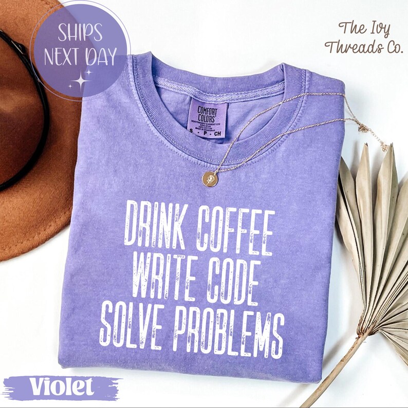 Peut inclure: Un t-shirt violet avec l'inscription blanche "DRINK COFFEE WRITE CODE SOLVE PROBLEMS". Un collier dor&eacute; et un chapeau marron sont visibles. Le t-shirt a une &eacute;tiquette Comfort Colors.