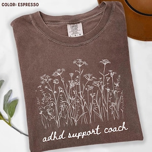 Puede incluir: Camiseta color espresso con cuello redondo y la etiqueta de la marca Comfort Colors. La camiseta presenta un diseño floral blanco y el texto "adhd support coach" en un estilo manuscrito. La camiseta está doblada sobre una superficie blanca.