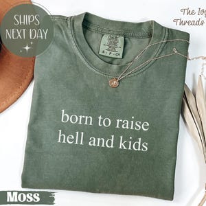 以下が含まれることがあります： モスグリーンのComfort Colors Tシャツ。白い文字で「born to raise hell and kids」と書かれています。金のネックレスがTシャツの上にあります。The Ivy Threads Co.のラベルが見えます。