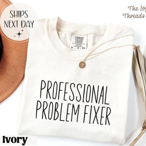 Può includere: T-shirt color avorio con la scritta "PROFESSIONAL PROBLEM FIXER" in nero. La maglietta ha girocollo e maniche corte. Una collana d'oro con un ciondolo è drappeggiata sulla maglietta. Il logo The Ivy Threads Co. si trova in alto a destra.