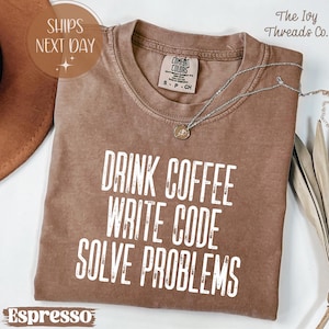 May include: Espresso-colored t-shirt with the text "DRINK COFFEE WRITE CODE SOLVE PROBLEMS" in white. The shirt has a crew neck and short sleeves. A silver necklace with a pendant is draped across the shirt.