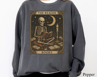 May include: Dark gray sweatshirt featuring a vintage-style graphic of a skeleton reading a book. The graphic includes the text "THE READER" and "BOOKS", with a crescent moon and candle. The sweatshirt is a comfortable, casual style.