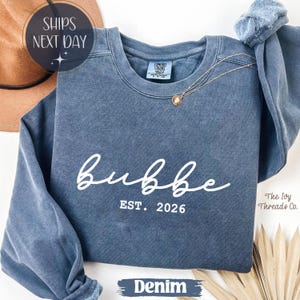 May include: A denim blue sweatshirt with the word "bubbe" in white script, and "EST. 2026" below. A gold necklace with a pendant rests on the shirt. The text "DENIM" is at the bottom.