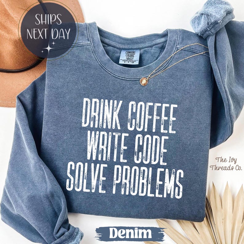 Peut inclure: Sweat-shirt bleu denim avec l'inscription blanche "DRINK COFFEE WRITE CODE SOLVE PROBLEMS". Le sweat-shirt a un col rond et des manches longues. Un collier en or avec un pendentif est drap&eacute; sur le v&ecirc;tement. Le logo The Ivy Threads Co. est en bas &agrave; droite.