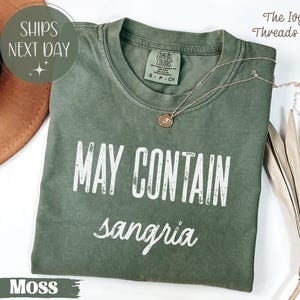 May include: Moss green t-shirt with the text "MAY CONTAIN sangria" in white. The shirt has a crew neck and short sleeves. A delicate gold necklace with a pendant rests on the shirt. The shirt is folded on a white surface.