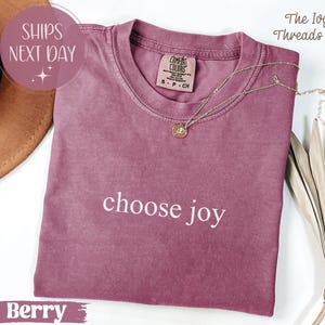 May include: Berry-colored t-shirt with the phrase "choose joy" in white lettering. A gold necklace with a pendant rests on the shirt. The shirt has a Comfort Colors tag. The text "SHIPS NEXT DAY" is in a pink circle.