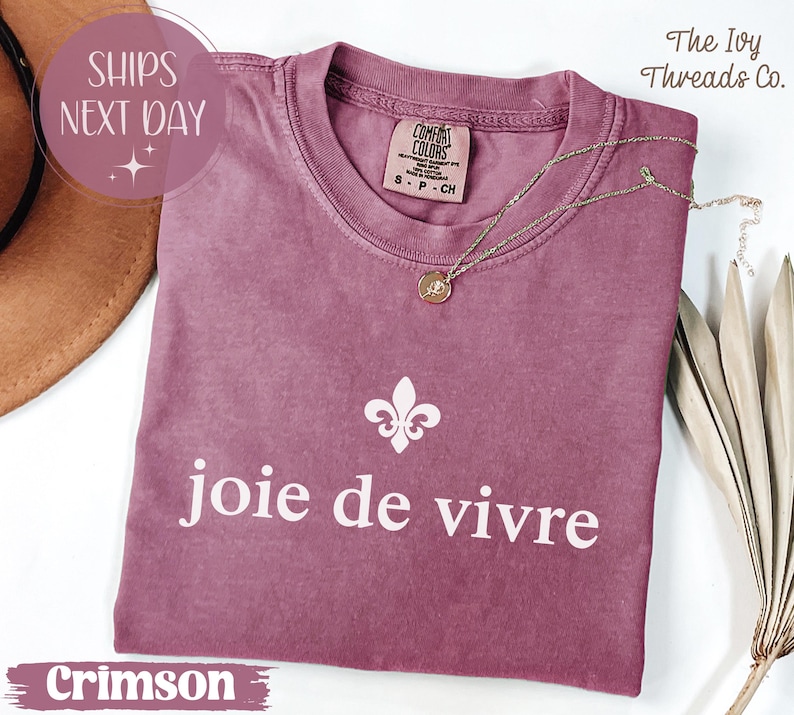 Puede incluir: Camiseta de color carmes&iacute; con la frase "joie de vivre" en blanco, un gr&aacute;fico de flor de lis y una etiqueta Comfort Colors. Un collar de oro con un colgante descansa sobre la camiseta. The Ivy Threads Co. est&aacute; impreso en la parte superior derecha.