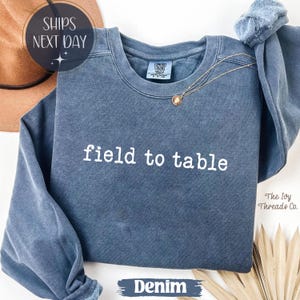 May include: Denim blue sweatshirt with the words "field to table" in white. A gold necklace with a pendant rests on the shirt. The text "The Ivy Threads Co." is visible on the right side of the image.
