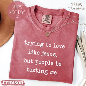 Peut inclure: T-shirt couleur cramoisi avec le texte "trying to love like jesus, but people be testing me." Le t-shirt a un col rond et une petite étiquette au dos. Un collier argenté avec un pendentif est drapé sur le t-shirt.
