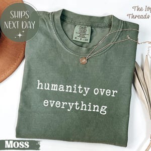 May include: A moss green t-shirt with the words "humanity over everything" in white. The shirt has a crew neck and short sleeves. A gold necklace with a pendant rests on the shirt. The Ivy Threads Co. is printed on the top right.