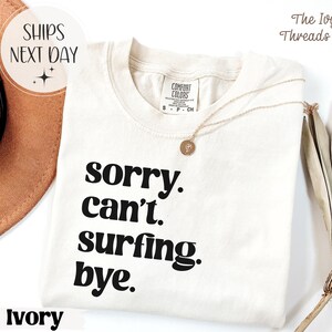 May include: Ivory-colored t-shirt with the text "sorry. can't. surfing. bye." in black. The shirt has a crew neck and short sleeves. A gold necklace with a pendant is draped on the shirt.