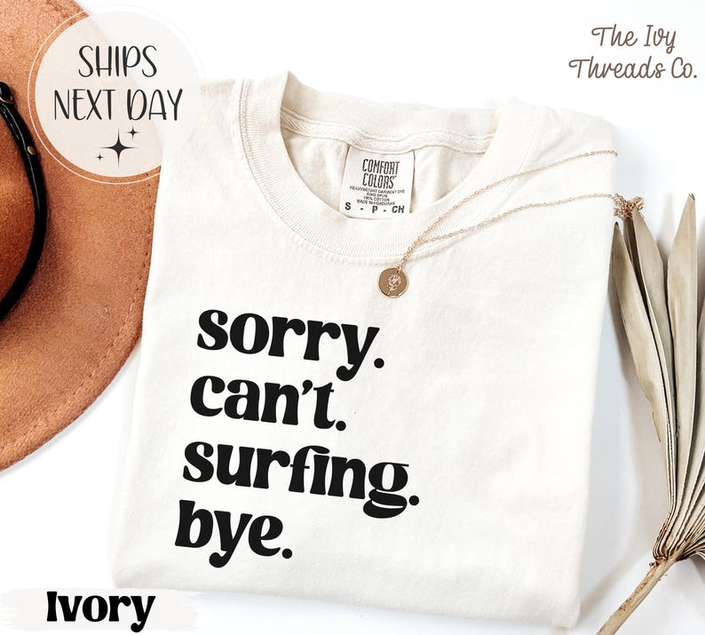 May include: Ivory-colored t-shirt with the text "sorry. can't. surfing. bye." in black. The shirt has a crew neck and short sleeves. A gold necklace with a pendant is draped on the shirt.