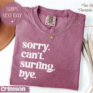 May include: Crimson-colored t-shirt with the text "sorry. can't. surfing. bye." in white. The shirt has a crew neck and a small tag on the collar. A gold necklace with a pendant is draped on the shirt.