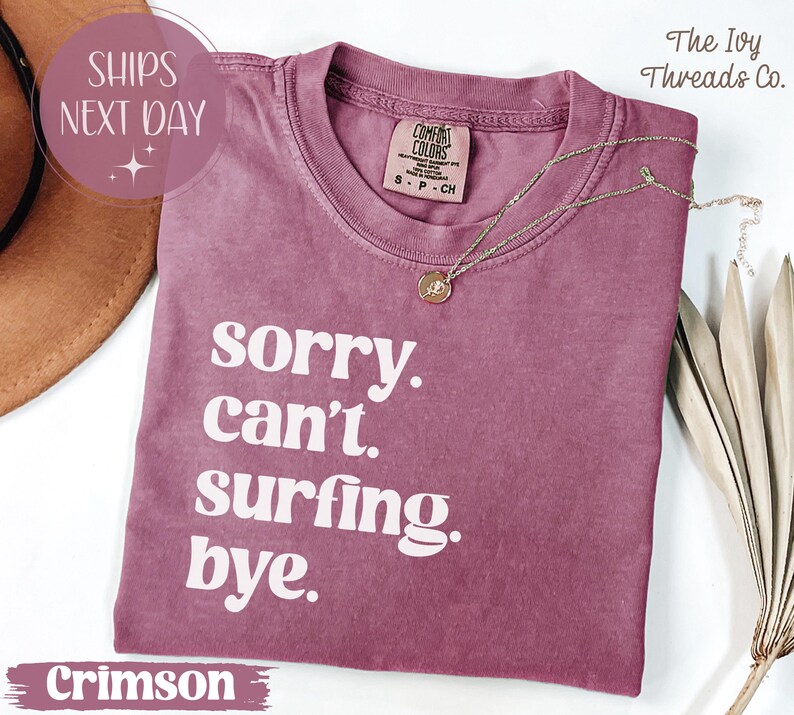 May include: Crimson-colored t-shirt with the text "sorry. can't. surfing. bye." in white. The shirt has a crew neck and a small tag on the collar. A gold necklace with a pendant is draped on the shirt.