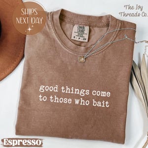 Könnte beinhalten: Espressofarbenes T-Shirt mit dem weißen Schriftzug „good things come to those who bait“. Eine silberne Halskette mit Anhänger liegt auf dem Shirt. Das Shirt ist mit „Comfort Colors“ gekennzeichnet und hat die Größe S.