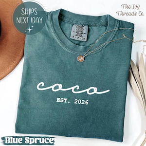 May include: A blue spruce Comfort Colors t-shirt with the word "coco" in white script and "EST. 2026" printed below. A gold necklace with a pendant rests on the shirt. The Ivy Threads Co. is printed on the top right.
