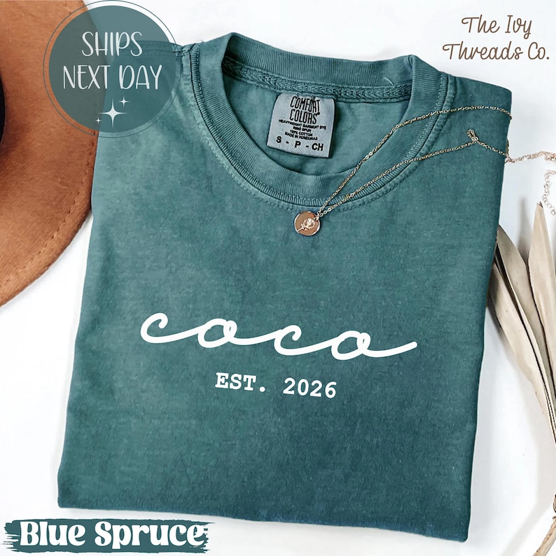 May include: A blue spruce Comfort Colors t-shirt with the word "coco" in white script and "EST. 2026" printed below. A gold necklace with a pendant rests on the shirt. The Ivy Threads Co. is printed on the top right.