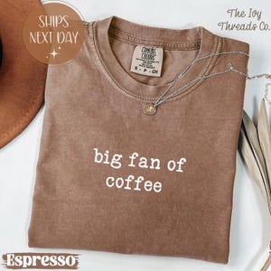 Könnte beinhalten: Espressofarbenes T-Shirt mit dem weißen Text "big fan of coffee". Das Shirt hat einen Rundhalsausschnitt und kurze Ärmel. Eine silberne Halskette mit Anhänger ist über dem Shirt drapiert. Das Shirt ist mit "Comfort Colors" gekennzeichnet.