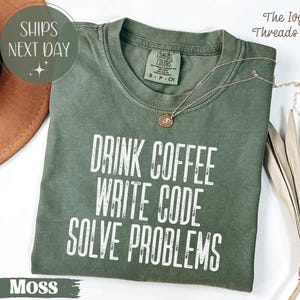 Peut inclure: T-shirt vert mousse avec l'inscription blanche &laquo; DRINK COFFEE WRITE CODE SOLVE PROBLEMS &raquo;. Le t-shirt a un col rond et une petite &eacute;tiquette au dos. Un collier dor&eacute; avec un pendentif est drap&eacute; sur le t-shirt.