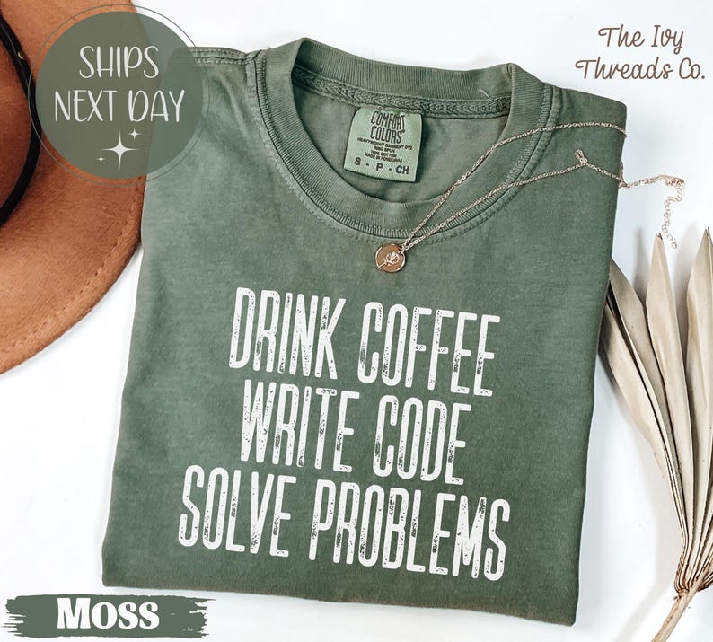 Peut inclure: T-shirt vert mousse avec l'inscription blanche &laquo; DRINK COFFEE WRITE CODE SOLVE PROBLEMS &raquo;. Le t-shirt a un col rond et une petite &eacute;tiquette au dos. Un collier dor&eacute; avec un pendentif est drap&eacute; sur le t-shirt.