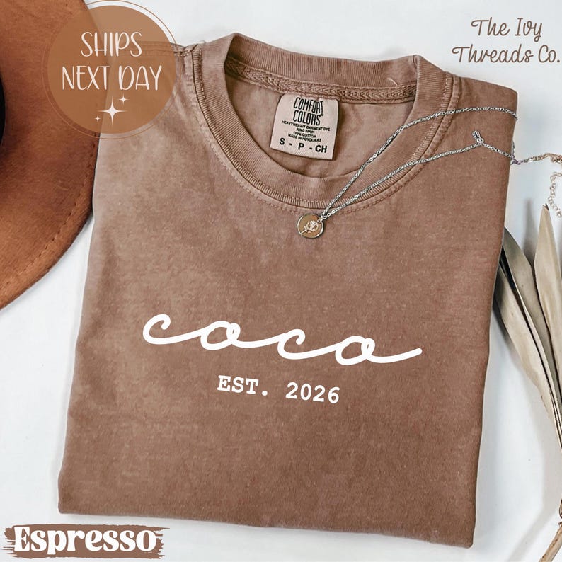 May include: Espresso-colored t-shirt with the word "coco" in white script and "EST. 2026" printed below. The shirt has a crew neck and a Comfort Colors tag. A silver necklace with a pendant is draped across the shirt.