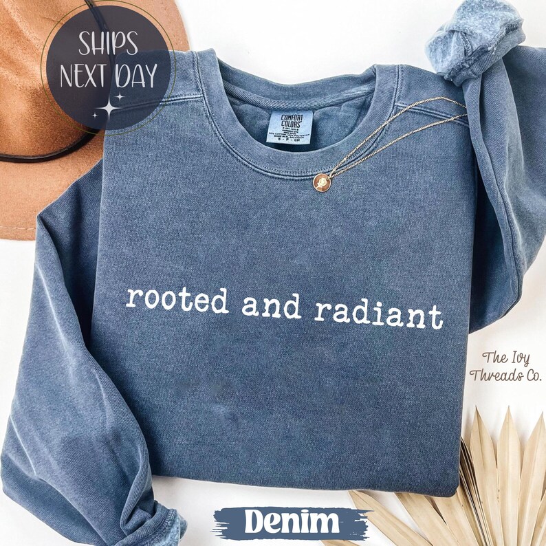 May include: Denim blue sweatshirt with the words "rooted and radiant" in white. A gold necklace with a pendant rests on the shirt. The sweatshirt has a crew neck and long sleeves. The text "Denim" is at the bottom.