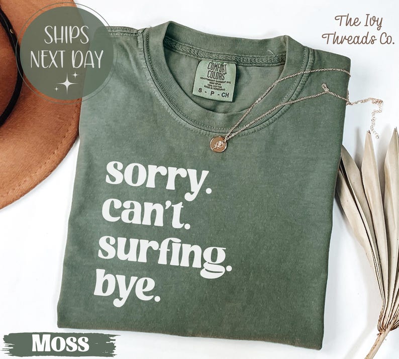 May include: Moss green t-shirt with the text "sorry. can't. surfing. bye." in white. The shirt has a crew neck and short sleeves. A gold necklace with a pendant is draped on the shirt.
