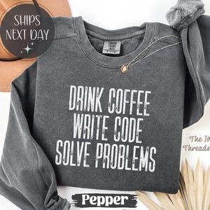 Peut inclure: Un sweat-shirt gris fonc&eacute; avec l'inscription blanche "DRINK COFFEE WRITE CODE SOLVE PROBLEMS". Le sweat-shirt a des manches longues et un col rond. Le mot "Pepper" est en bas de l'image.